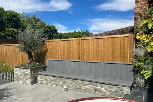 Long stone-faced bench with back and seat of Brushed Basalt Millboard decking boards cut to size, on patio, against wooden fence.