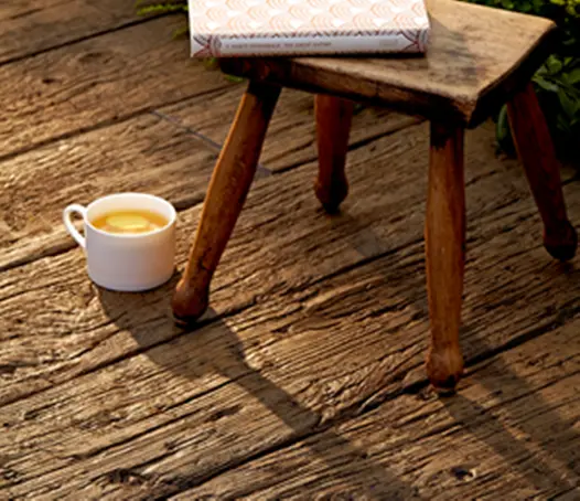 Close-up of Vintage Millboard Decking with a chair, book, and latte, emphasizing the cosy, timeless appeal of the weathered wood grain.