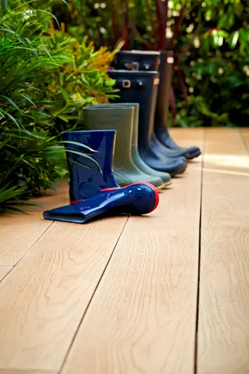 Four sets of wellington boots sit on golden oak millboard decking next to green hedge, one boot is on its side.