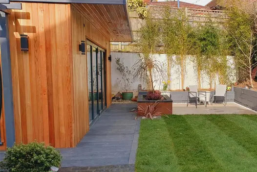Red wood clad building leads out to small burnt cedar decking area with corten steel planters and lawn separating patio area.