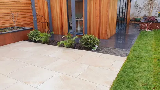 Ash Beige porcelain patio in front of a wooden cladded garden building, a raised flower bed on one side and a lawn on the other side.