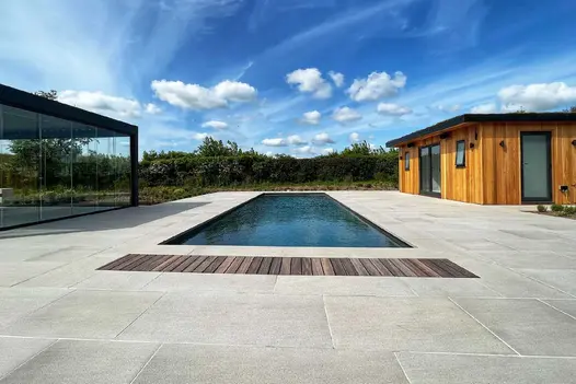 Urban Grey Porcelain paving used in large paved area with pool in the middle. Glass door pergola on the left with an outhouse on the right.
