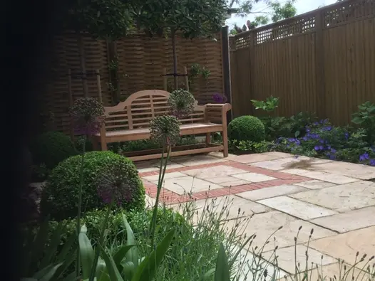 Lutyens-style bench at one end of Mint Indian sandstone paving with brick detail, next to border below tall fence.