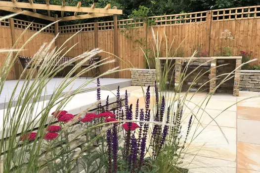 Corner of fenced patio with brick barbecue on Mint Indian sandstone paving, next to composite decking with pergola and seats.