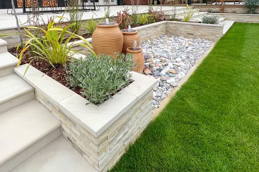 Patio with 2 sets of steps descending either side of pebble area enclosed on 3 sides by raised bed with Mint sandstone cladding.