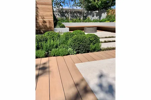 Row of hedges line edge of mocha designboard composite decking leading to indented patio with built in bench and fire pit.
