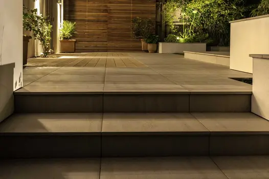 Views at night show 2 steps lead up to patio and decking area with light tiles and mocha designboard composite decking.