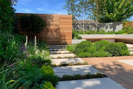 Trees cast shadows on mocha designboard decking in garden filled with foliage, built in corner bench and fire pit in the background.