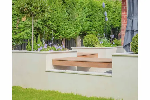 WPC decking Mocha Designboard fitted as bench between cream walls edging raised beds at side of patio. Leafy trees in background.