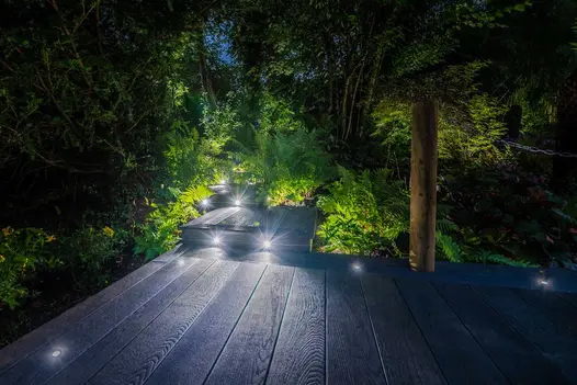 Night view of woodland borders on either side of staggered sections of Burnt Cedar Millboard decking with inset light spots.