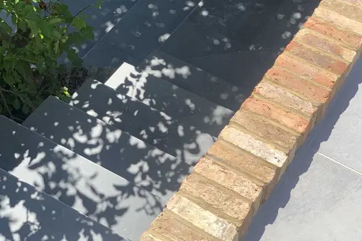 Dappled shadow plays on 6 Brazilian Black slate steps descending past brick wall to paving below. Built by Monarch.