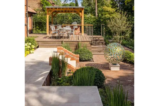 Patio of pergola-covered decking and Egyptian Beige limestone on 2 levels with steps down to clay paving with ball sculpture.