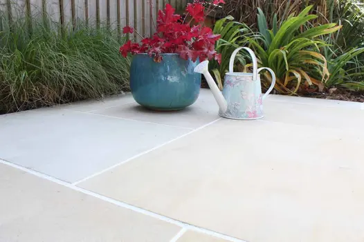 Rust-coloured heuchera in blue pot sits by miniature watering can on Multi Mint smooth paving by planted beds.
