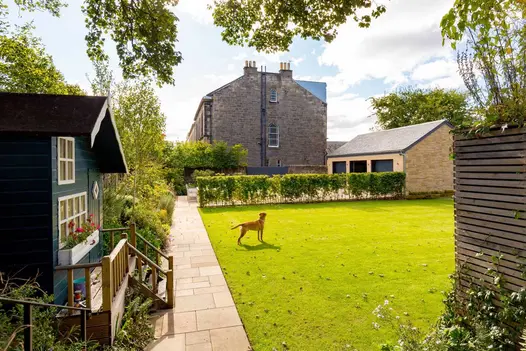 Buff smooth sandstone path runs between lawn and bed with large Wendy house. Dog stands on grass. Design by Murray Developments.