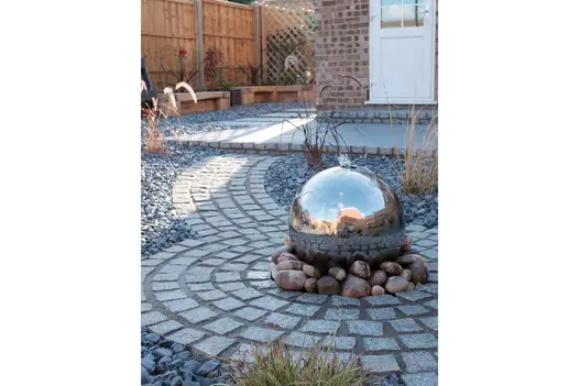 Gravel garden with curved path and circle of silver grey granite setts, silver ball fountain, grasses and raised wooden beds.