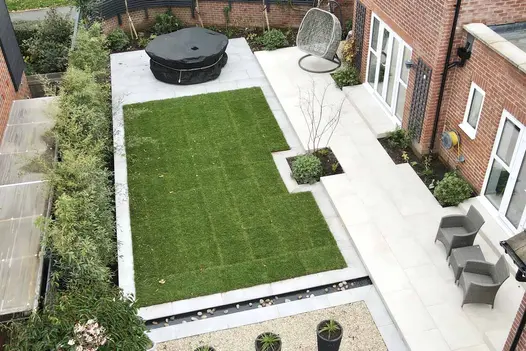 View down on rectangular lawn next to long patio of Florence Beige running along back of house. Covered hot tub and planted borders.