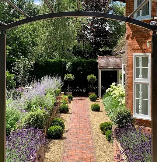 Narrow path of Dorset Antique Clay Pavers runs through pebbles, with house on the right and huge trees and lavender plants to the left.