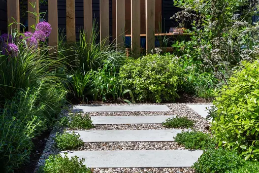 Long planks of Jura Grey sawn limestone paving set into gravel, bordered on 2 sides by beds planted with grasses and low shrubs.