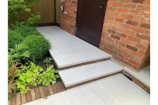 Two Faro porcelain 40mm downstand steps leading up to small patio next to brick outbuilding with black door.