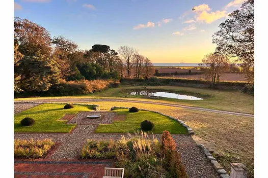 Old English clay pavers edge geometrically shaped lawn with water bowl in central paved square. View to horizon across fields.
