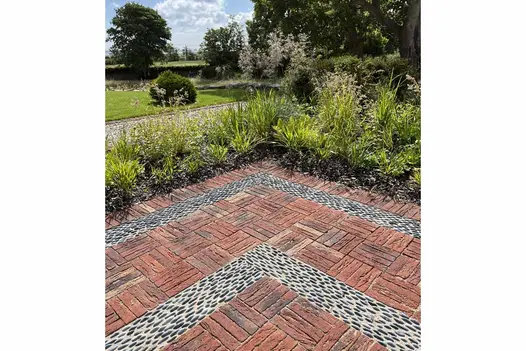 Planted border edges corner of paved area of Old English clay bricks laid in basketweave pattern with strips of pebbles inlaid. 