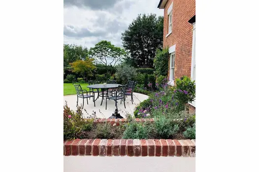 White rendered raised bed with Old English clay pavers as coping. Round patio with metal dining set beyond. 