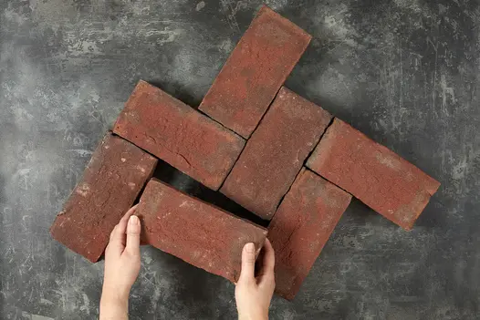 2 hands place 7th Old English clay paver into formation of 6 matching bricks against dark background. Free UK delivery available.