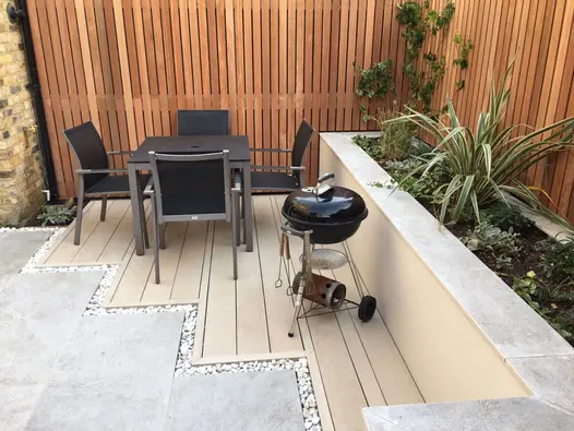 Small, unique decking area of golden oak millboard hosts quaint dining table and BBQ next to red cedar fencing.