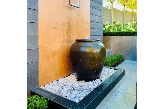 Water feature using dark plant pout has flows coming out the top, falling on to light pebbles, frame clad with opium black designclad.