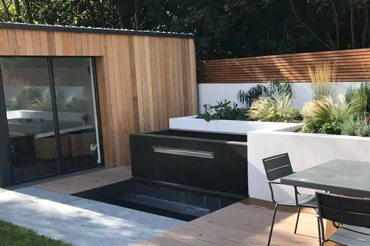 White rendered walls with built in flowerbeds has opium black designclad waterfeature next to large outdoor building.