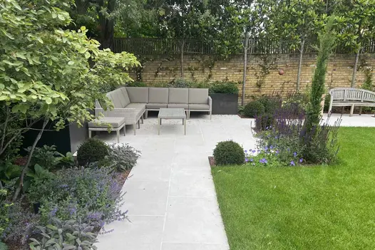 Florence White porcelain path, 2 slabs wide, leads to square paved area in corner of garden with modular sofa and table.