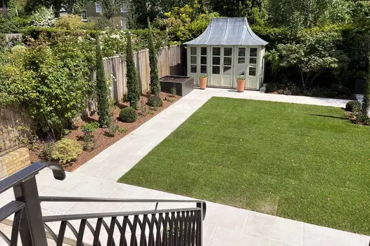 View down ornate metal hand rail towards summer house at back of garden with square lawn edged with Florence White porcelain path.