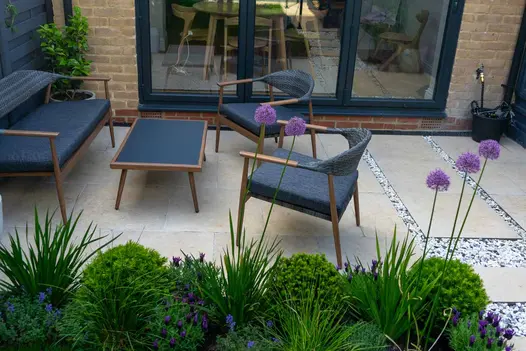 Patio of Jura Beige porcelain paving with modern outdoor furniture sits outside 3-panel bifold doors of brick house.