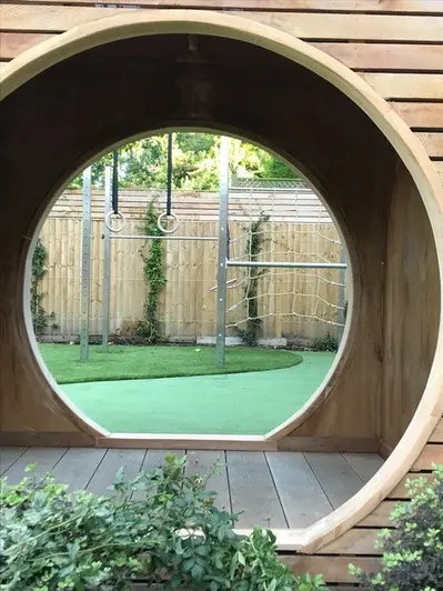 Metal climbing frame with ropes and swings seen through circular tunnel with golden oak millboard decking laid inside.