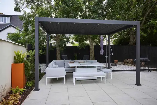 String lights adorn the proteus grey weatherproof pergola, with garden furniture sitting on astor grey porcelain paving.