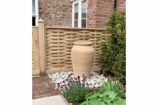 Old English clay pavers laid in front of garden gate in wooden fence next to tall ribbed pot on white pebbles.