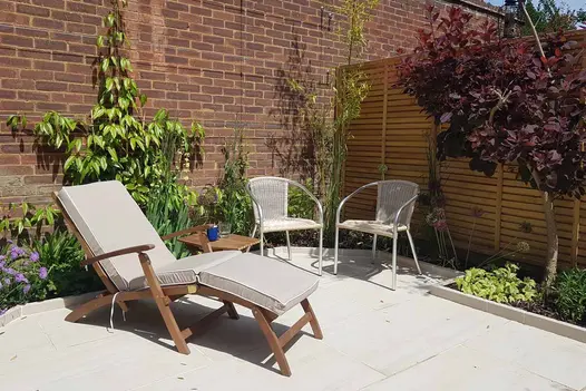 Sun lounger sits on Faro Porcelain Paving next to two rattan armchairs with table in between holding drinks. 
