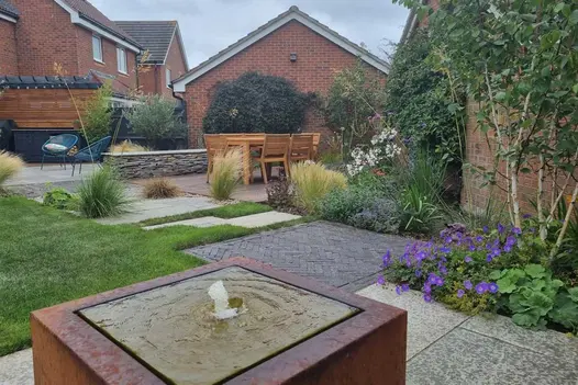 Garden with water feature and furniture on mixed surfaces of Kandla Grey Indian sandstone paving, clay pavers, lawn, and gravel.