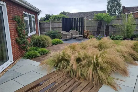 Back garden of surburban house with Kota Blue limestone paving laid with decking, punctuated with small lawn and grassy planting.