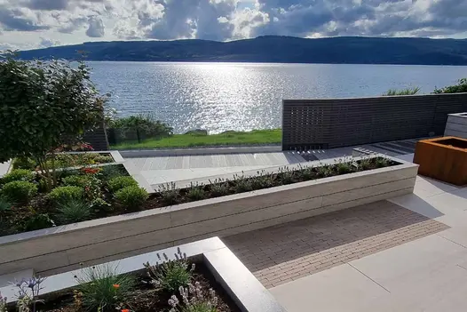 Composite raised planters clad in pebble grey composite battens overlooks glistening lake with mountains in the distance.