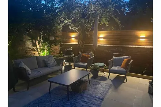 Seen at night, this impressive view shows off lounge area with sofa and chairs surrounding a table on top of harvest smooth sandstone paving.