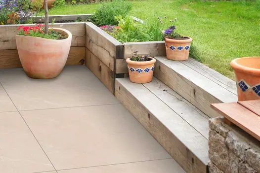Garden patio paved with Piazza Beige porcelain, styled with rustic timber sleepers and terracotta planters.