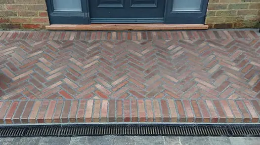 Wide platform step of mixed Winton and Bexhill clay pavers, channel drain at base, lies beneath blue front door. 