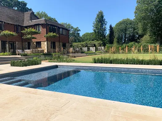 Rectangular blue swimming pool set into Jura Beige sawn limestone paving in large garden with lawn and patio across back of house.