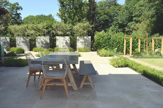 Long patio of Jura Beige limestone sawn paving with dining set, planted borders and, at far end, a hedge punctuated by screens.