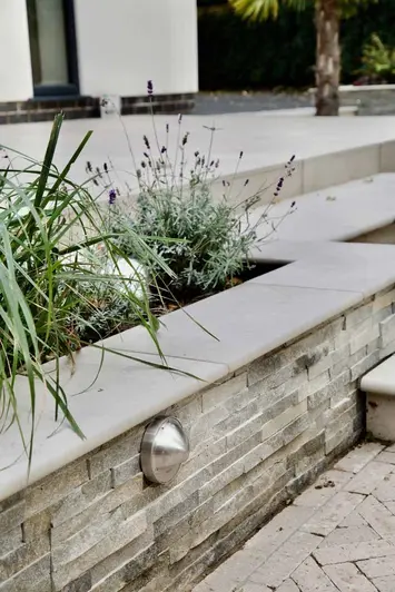 Close up of raised flowerbed clad with stone walling and capped with ash beige porcelain 20mm bullnose coping stones.