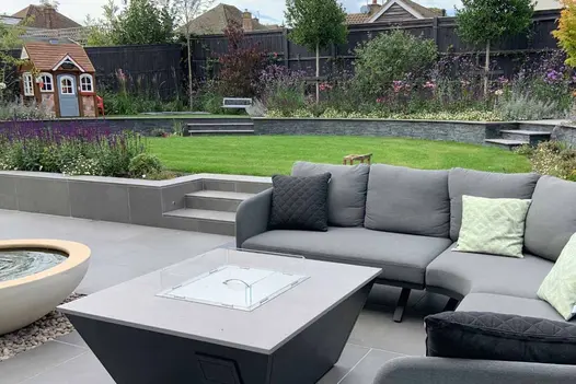 Fire-pit around grey garden furniture with steel grey porcelain garden steps leading up to lawn, level up shows a childrens play area.
