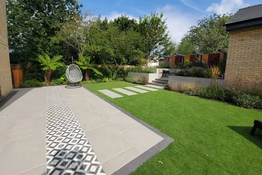 Egg chair sits by tree fern at far end of patio of decorative tiles and porcelain slabs, edged with Platinum Grey Plank Paving.