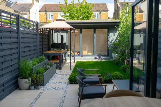 Path of Jura Beige porcelain outdoor tiles UK set into gravel leads from patio area to garden studio past a covered work bench.