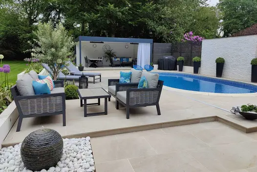 Swimming pool terrace with oval pool, seating area, sun loungers and open sided pergola. Paved with rectangle porcelain paving and bullnose steps.